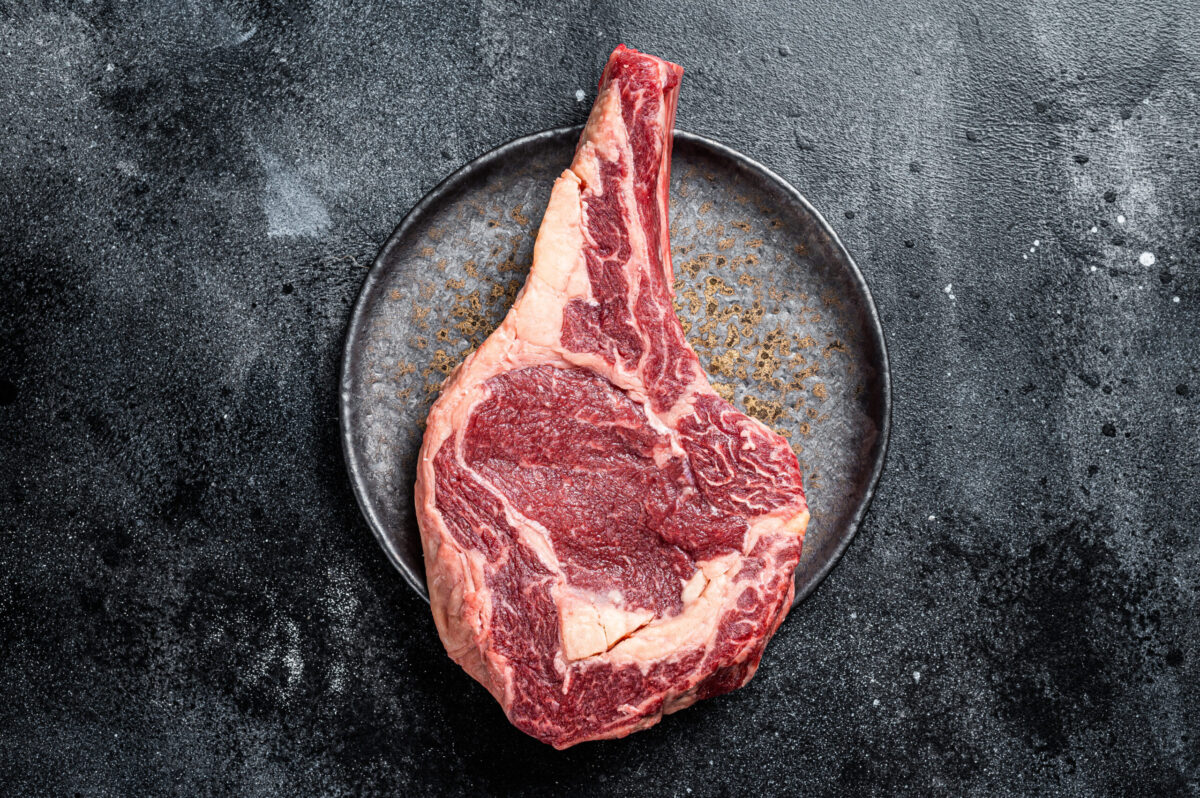 Raw cowboy steak, rib eye steak with bone, beef marbled meat on butcher cutting board. Black background. Top view. Copy space.